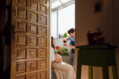 Sakallı genç Latin beyaz adam içki arkadaşı ile evde konuşuyor, ön kapıdan evin içinden görülüyor..