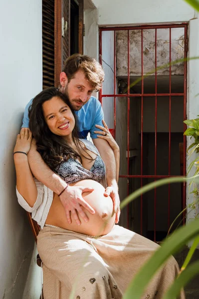 妊娠中の妻を家で抱き込み笑顔でカメラを見ている若いラテン系のカップルの縦の肖像画 — ストック写真