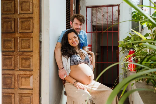 妊娠中のラテン語の若いカップルの肖像画家を見てカメラと笑顔 アルゼンチン人男性と彼のブラジル人の妻と一緒に家に座っています — ストック写真