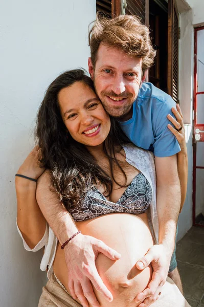 髭を生やした若いアルゼンチン人の白人男性が妊娠中のブラジル人の妻を抱きかかえ笑顔でカメラを見ています — ストック写真