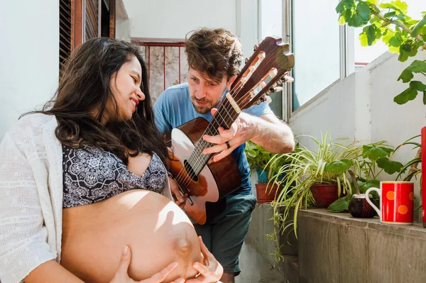 若いロマンチックな妊婦ラテンカップル白人のアルゼンチン人男性彼のブラジル人の妻と一緒に音楽を作って家でリラックスした歌を楽しんでいます — ストック写真