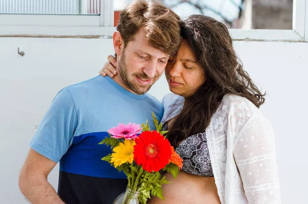 妊娠中の若い夫婦のアルゼンチン人男性とブラジル人女性はお互いの花を抱き合って家に立っていて花を見ていて — ストック写真