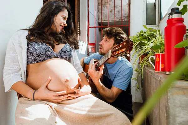 ブラジルの民族の妊婦は家に座って彼女の夫が彼女のためにギターを演奏し 再び恋に落ちるのを見て 一緒に新しい赤ちゃんを待っている家族として楽しんでいます — ストック写真
