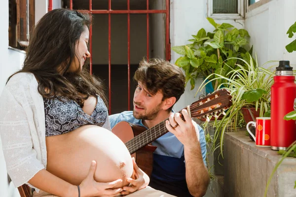 自宅で髭を生やした若いラテン系のアルゼンチン人男性がギターを弾いている間に間違いを犯し妻は若い妊婦のブラジル人女性が彼女の腹に触れる椅子に座って彼を見て笑います — ストック写真