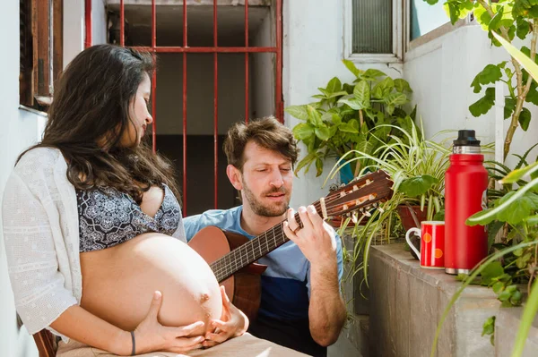 短い髪とひげを持つ若いアルゼンチン人男性が家でうずくまっていて 妊娠しているブラジルの民族の女性にギターを演奏し 彼女の腹に座って触れている方法を示しています — ストック写真