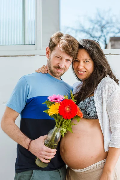 彼のブラジル人の妻とアルゼンチン人男性の成人妊娠ラテンカップルのウエストアップ肖像画 屋内でカメラを見て立って ガラス花瓶の中にある花を保持 — ストック写真