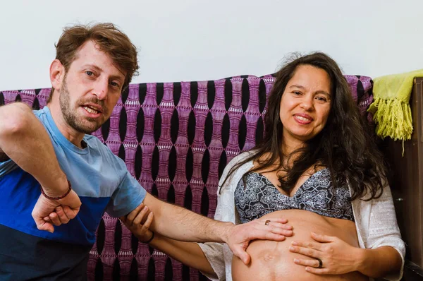 髭を生やした白人の成人男性がカメラを見ながら家に座って子供が妊娠中のブラジル人女性の子宮の中でどのように動くかを説明しました — ストック写真
