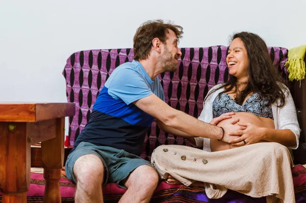 一对拉丁裔阿根廷成年夫妇和他的巴西妻子坐在沙发上 他们兴奋地微笑着 感受着未来女儿在子宫里的动作 — 图库照片