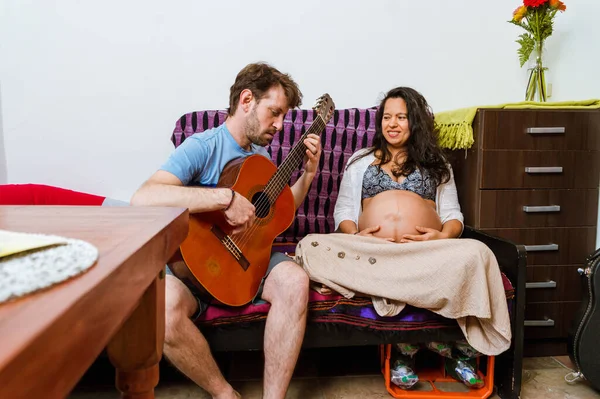 アルゼンチン人のひげを生やした白人の若い大人の男 彼の妊娠中の妻ブラジル人の大人の女性を楽しませます 彼女のために自宅でギターを演奏 リビングルームのソファに座って 楽しい家族の時間を持っています — ストック写真