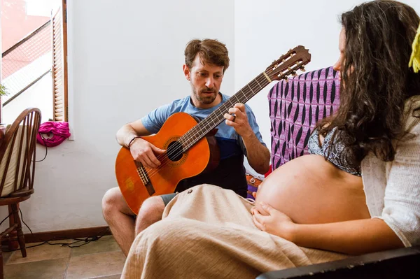 romantic young man plays guitar to his pregnant wife a young brazilian woman, sitting on the sofa inside the house