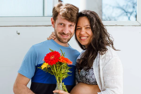 妊娠中のラテン系のカップルの肖像画を家に持ち花を持ち抱きかかえてカメラを見て笑っていました — ストック写真