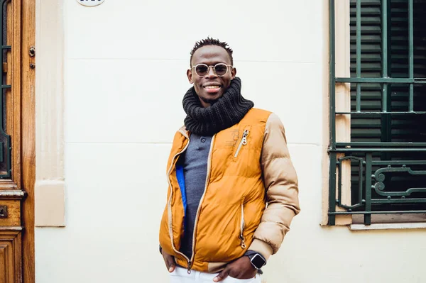 happy african descent man laughing looking at the camera posing in an urban street, wearing glasses, scarf and winter clothes, standing between a wall and a window.