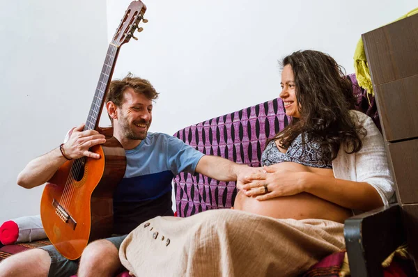 白人成年男子一边在吉他上弹出一首歌 一边坐在屋里的沙发上 高兴地微笑着 触摸着妻子的腹部 感受着胎儿在子宫里的运动 — 图库照片