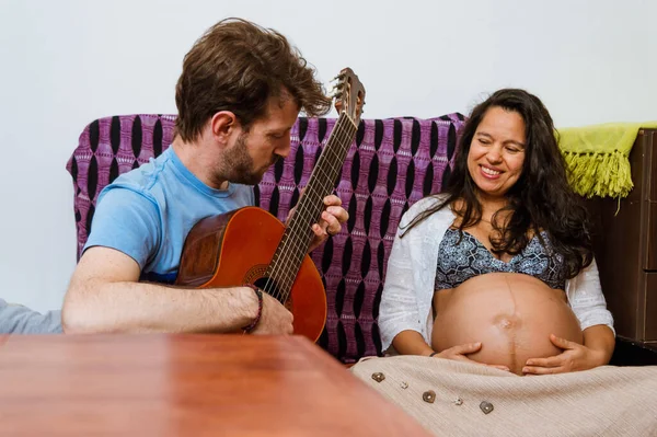 彼の妻の子宮の中にある彼の赤ちゃんのためにアコースティックギターを演奏大人の白人のアルゼンチン人男性 ブルネットのブラジル人の大人の女性 家の中のソファに座って一緒に楽しみます — ストック写真