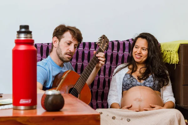 アルゼンチンの白人成人男性は家の中でギターを弾くことに触発され 妻はブラジル人の妊婦が彼と笑顔を見て 彼の音楽を楽しんで ソファに座っています — ストック写真