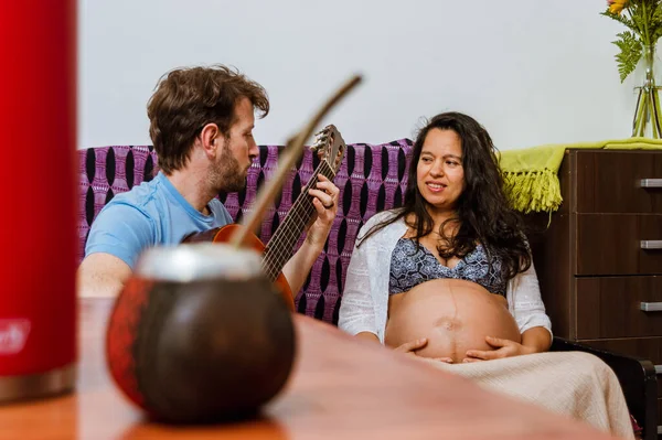 妊娠中のブラジル人女性彼女の夫を見てアルゼンチン人の若い大人がギターを弾きます 家の中のアームチェアに座っています 一緒に週末を楽しんで — ストック写真