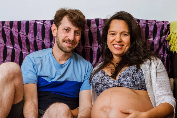 髭を生やした白人のアルゼンチン人男性と妊娠中のブラジル人の妻がリビングのソファに座ってカメラを見て笑っていました — ストック写真