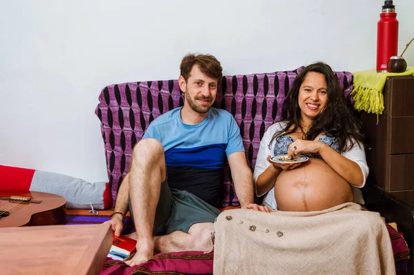 妊娠中の妻の隣に座っている白人の大人の男が休憩室でブラジル人の女性でカメラを見て笑っています — ストック写真