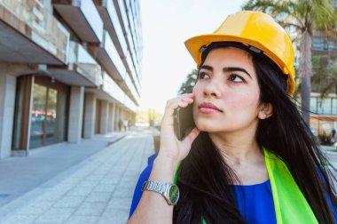 Beyaz kadın mühendis, güvenlik kaskı ve yeleği takıyor, binanın yanında dikiliyor, telefonla konuşuyor ve yan tarafa bakıyor..