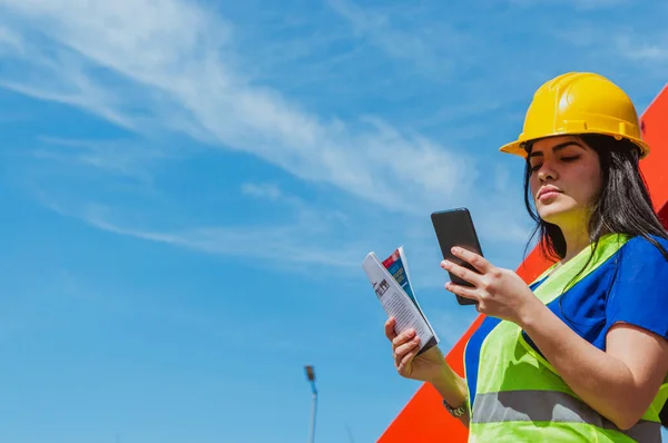 young latin caucasian outdoor worker woman checking notifications on her phone, engineering and industry concept, copy space.