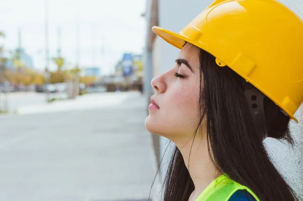 young caucasian engineer woman wearing yellow protective helmet, exhausted from work, resting leaning against a wall with her eyes closed, copy space, industry concept.