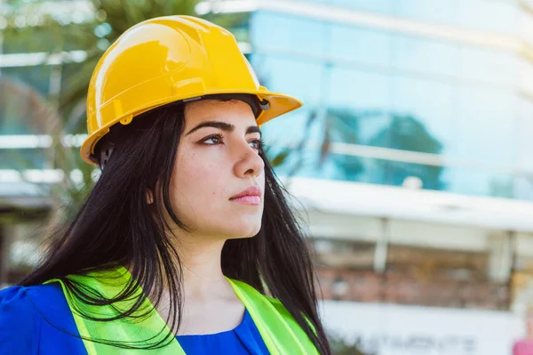 portrait of young caucasian female engineer, dressed in blue with yellow protective helmet and safety vest, standing outdoors looking into the distance thinking, copy space, industry concept.