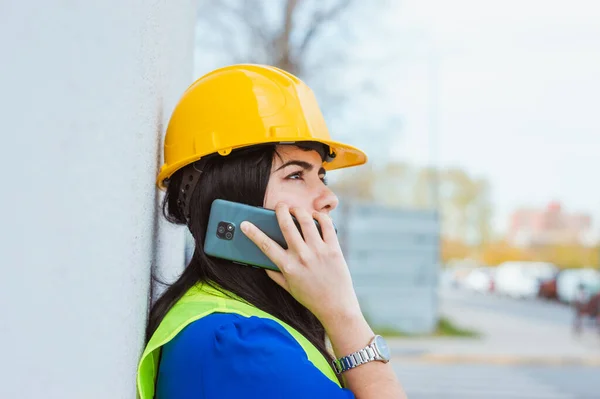 young latin woman engineer with safety helmet and vest, standing outdoors, leaning against a wall, she is on a call talking to her boss on the phone and looking around, copy space, industry concept.