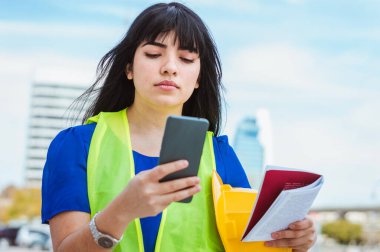 Genç beyaz Venezuelalı kadın, mühendis, elinde telefonuyla caddede duruyor, patronundan gelen mesajı dikkatle okuyor, bildirimleri ve sosyal ağları kontrol ediyor.