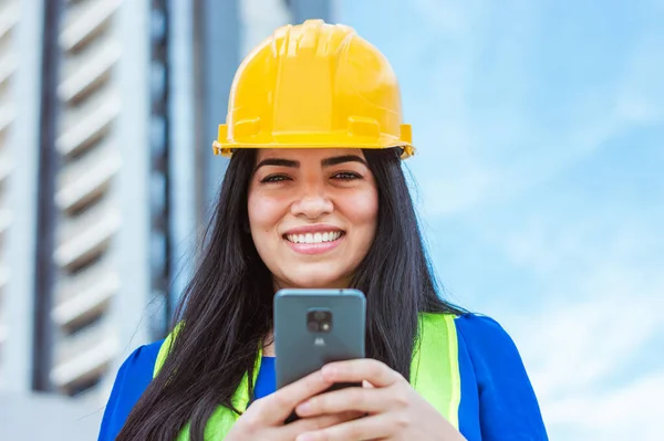 Caucasian young woman, engineering professional wears a yellow helmet, reflective vest, outdoors looking at the camera, smiling and holding a phone, with buildings and the sky in the background.