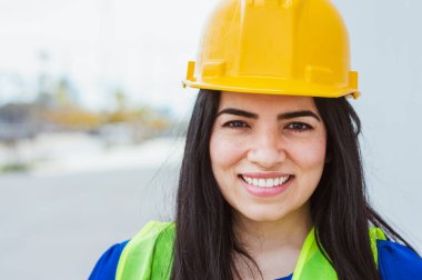 Güvenlik kaskı ve yelek giyen genç Latin kadın mühendis, inşaat alanında çalışan, caddede durup kameraya gülümseyen, fotokopi alanı, inşaat ve endüstri konseptine bakan.