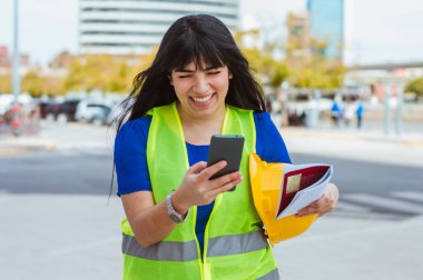 Beyaz tenli genç kadın mühendis sokakta durmuş gülümsüyor sosyal medya, endüstri ve teknoloji konseptindeki mesaj ve bildirimleri okuyor..