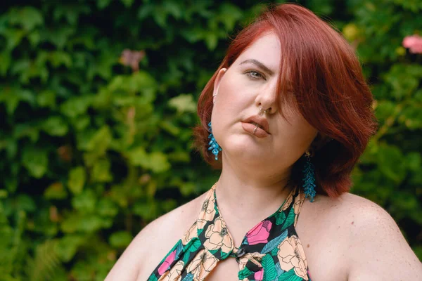 portrait of beautiful young latina caucasian plus size model woman, short red hair, dressed in a flowered dress, relaxed and calm standing in a garden looking at the camera. copy space.