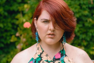 Beyaz Latin kökenli, kısa kızıl saçlı bir kadının portresi, artı boyut modeli, kameraya ciddi bir şekilde bakıyor, arka planda bitki örtüsüyle dışarıda duruyor..