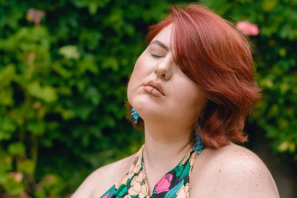 young latina caucasian plus size model woman with short red hair standing outdoors with eyes closed, relaxed, inspired, thinking and breathing, with copy space and greenery in the background.