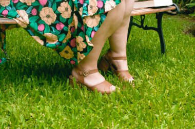 Tanımlanamayan beyaz kadın bacakları, artı beden modeli, bir bahçede ahşap bir bankta oturuyor, çiçekli bir elbise giyiyor. Boşluğu kopyala.