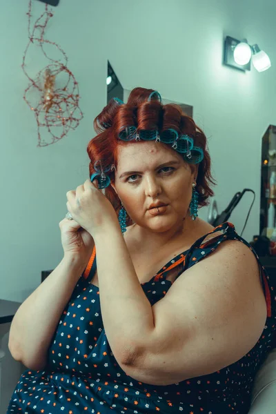 Caucasian young woman of Argentinian, Hispanic Italian ethnicity, red hair, sitting at hair salon putting curlers together with rollers, looking at camera