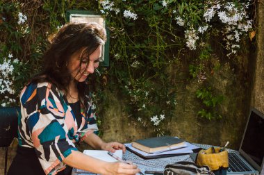 Fransız kökenli güzel, mutlu, beyaz bir kadın müşterisi için tasarım yaparken gülümsüyor, evinin terasında dizüstü bilgisayarıyla oturmuş, kağıda çizimler yapıyor..