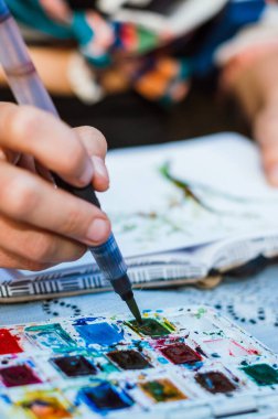 closeup vertical image of unrecognizable caucasian woman dipping the brush in watercolor, making a drawing, sitting on the terrace of the house during sunset.