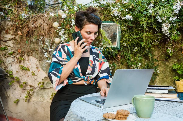 Caucasian adult woman of French ethnicity, working on the terrace with her laptop and listening to a voice memo from her phone, with a cup of coffee and cookies on the table, copy space.