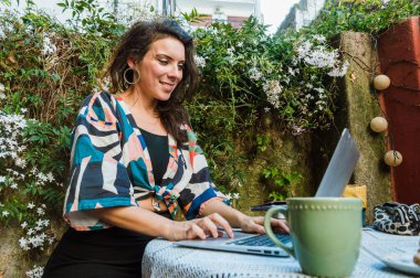 Happy excited caucasian adult woman watching laptop, surfing the internet, checking emails and notifications, working sitting on the terrace, with a cup of coffee on the table.