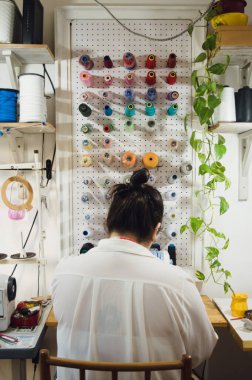 rear view of young woman sitting in her sewing workshop working with the sewing machine, with coils of thread arranged on the background wall.