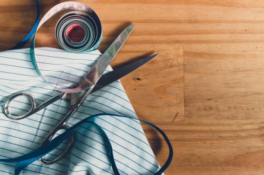 top view of steel scissors on a white cloth with blue stripes and measuring tape placed on top of a wooden table, with copy space on the right.