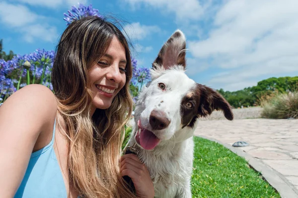 portrait of border collie dog with white fur with brown and green eyes looking at the camera with a funny face next to his owner who is happy smiling and looking at him, with sky on background.