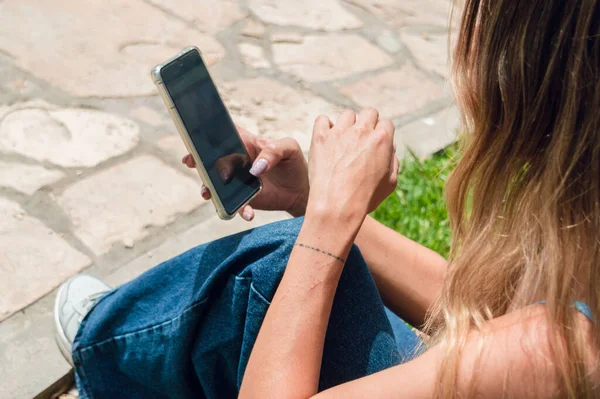 rear view of young woman with long hair and blue clothes, sitting outdoors, in a park, checking social networks on her phone, watching the screen, communication concept, copy space.