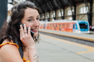 beautiful young latin woman of argentinian ethnicity, on a phone call smiling and looking at the camera sitting waiting for the train on the platform, travel and technology concept, copy space.