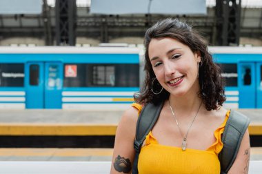 beautiful young latin woman of argentinian ethnicity, dressed in yellow, is sitting on the platform, looking at the camera and smiling happy because she is going on a train trip through Buenos Aires