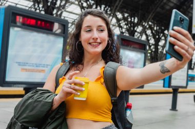 young latin woman of argentinian ethnicity, taking a selfie, sending a photo message on her phone while waiting for the train on the platform.