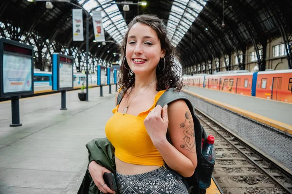 portrait of young Latina woman of Argentinian ethnicity with white skin, with a tattoo on her arm, dressed in yellow and with a backpack, standing on the train platform, smiling looking at the camera