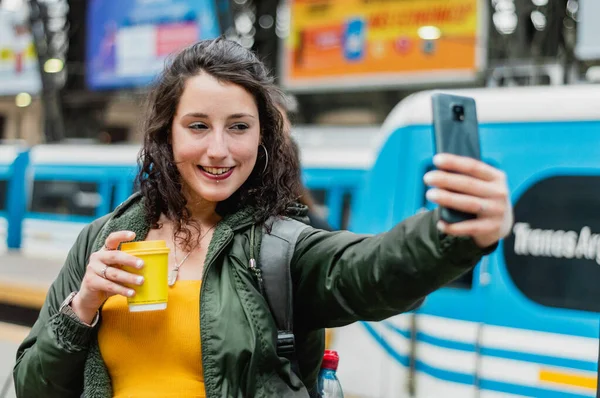 young latin woman from Argentina, standing on the platform taking a selfie with her phone, happy smiling because she is going on a train trip in the province of Buenos Aires