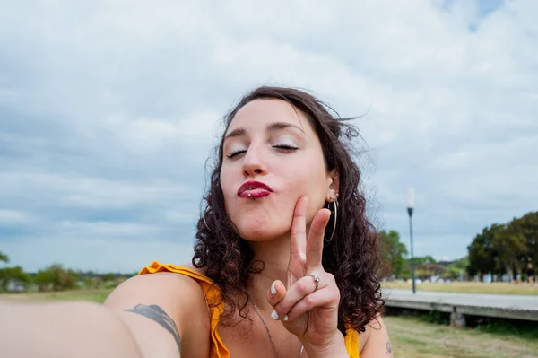 Selfie portrait of young Argentinian Latina woman, white-skinned with red lips and dressed in yellow, outdoors looking at the camera giving a kiss and making a peace and love sign, lifestyle concept.